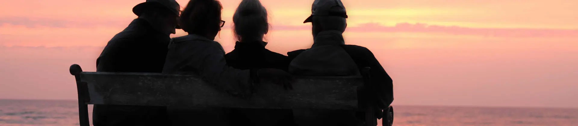 Over 70s friends sitting on a bench looking out at the sea at sunset