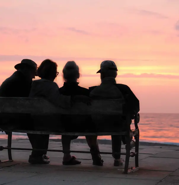 Over 70s friends sitting on a bench looking out at the sea at sunset