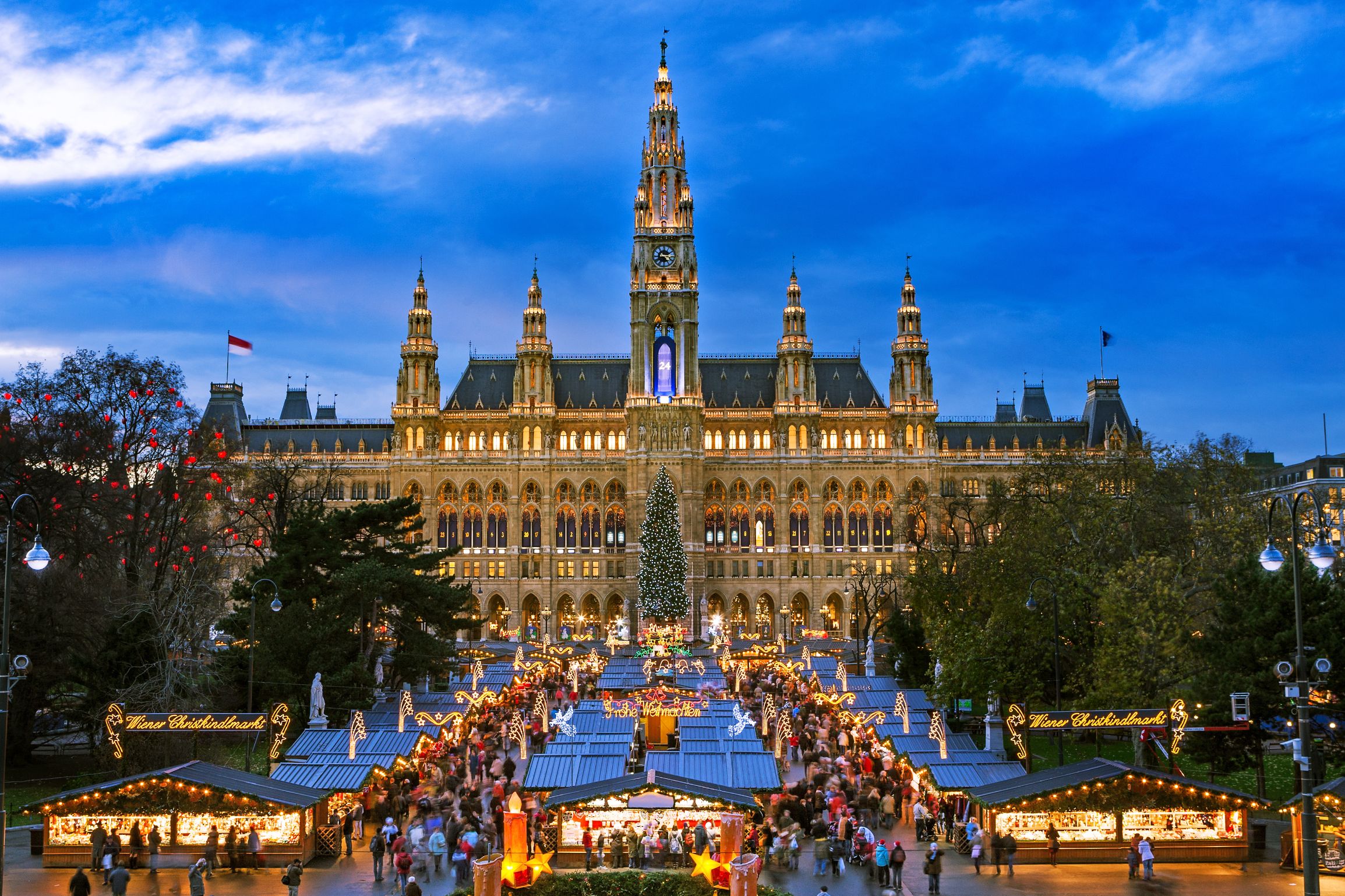 Shot of a gothic city hall which has a tall clock tower in the middle and four smaller towers, two either side, all with spiky turrets. The building has long windows all over and is a gold colour. Below are the Vienna Christmas markets, with four vertical strips of stalls, the centre having a christmas tree at the far end. There are many people shopping, with lights and lit up signs above them.