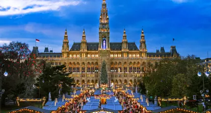 Shot of a gothic city hall which has a tall clock tower in the middle and four smaller towers, two either side, all with spiky turrets. The building has long windows all over and is a gold colour. Below are the Vienna Christmas markets, with four vertical strips of stalls, the centre having a christmas tree at the far end. There are many people shopping, with lights and lit up signs above them.