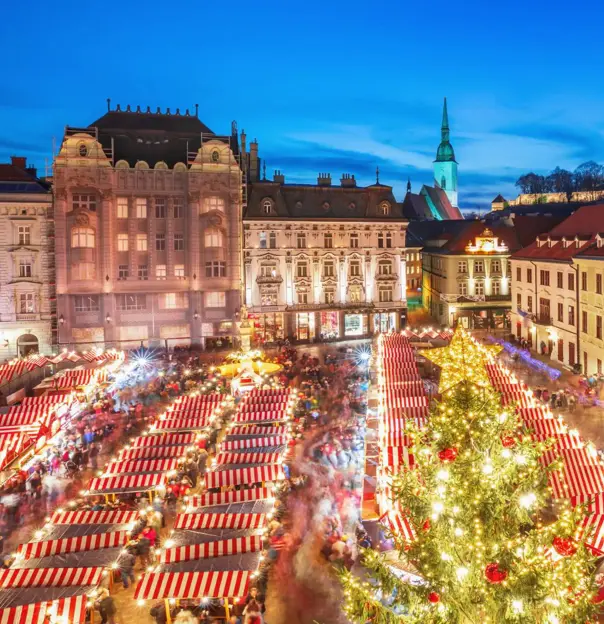 A Christmas market in Bratislava's main square, featuring festive wooden stalls decorated with lights, a large Christmas tree, and crowds of people enjoying the festive atmosphere
