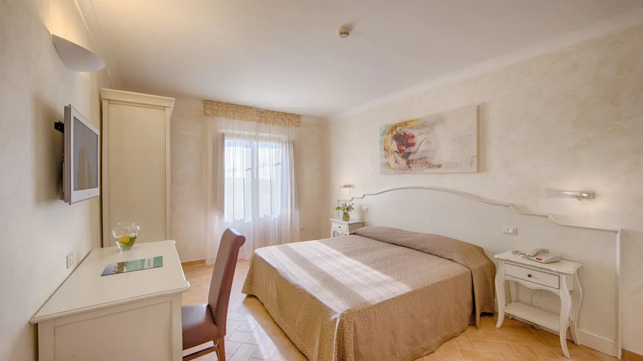 A bedroom at Masseria Casselli Hotel with a double bed covered in a brown bedspread, white bedside tables, a desk with a chair, wall-mounted TV, and a window with sheer curtains letting in natural light