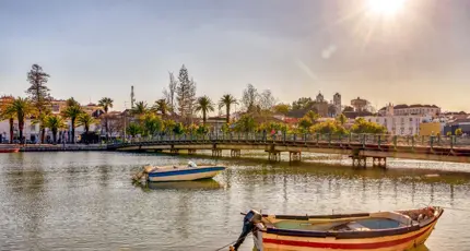 Two small boats moored in front of a bridge in Tavira on the Algarve, with trees and buildings lining the water’s edge in the background