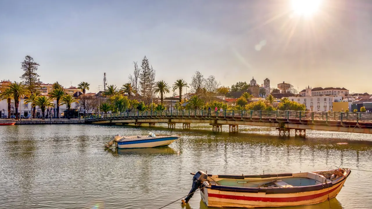 Two small boats moored in front of a bridge in Tavira on the Algarve, with trees and buildings lining the water’s edge in the background