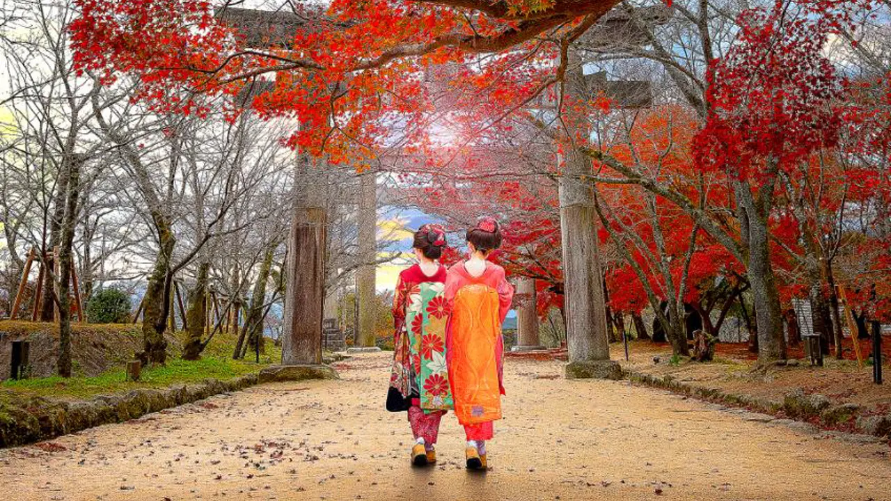 Two women in traditional kimono walk beneath vibrant autumn leaves toward a torii gate at Homangu Kamado Shrine in Fukuoka, Japan