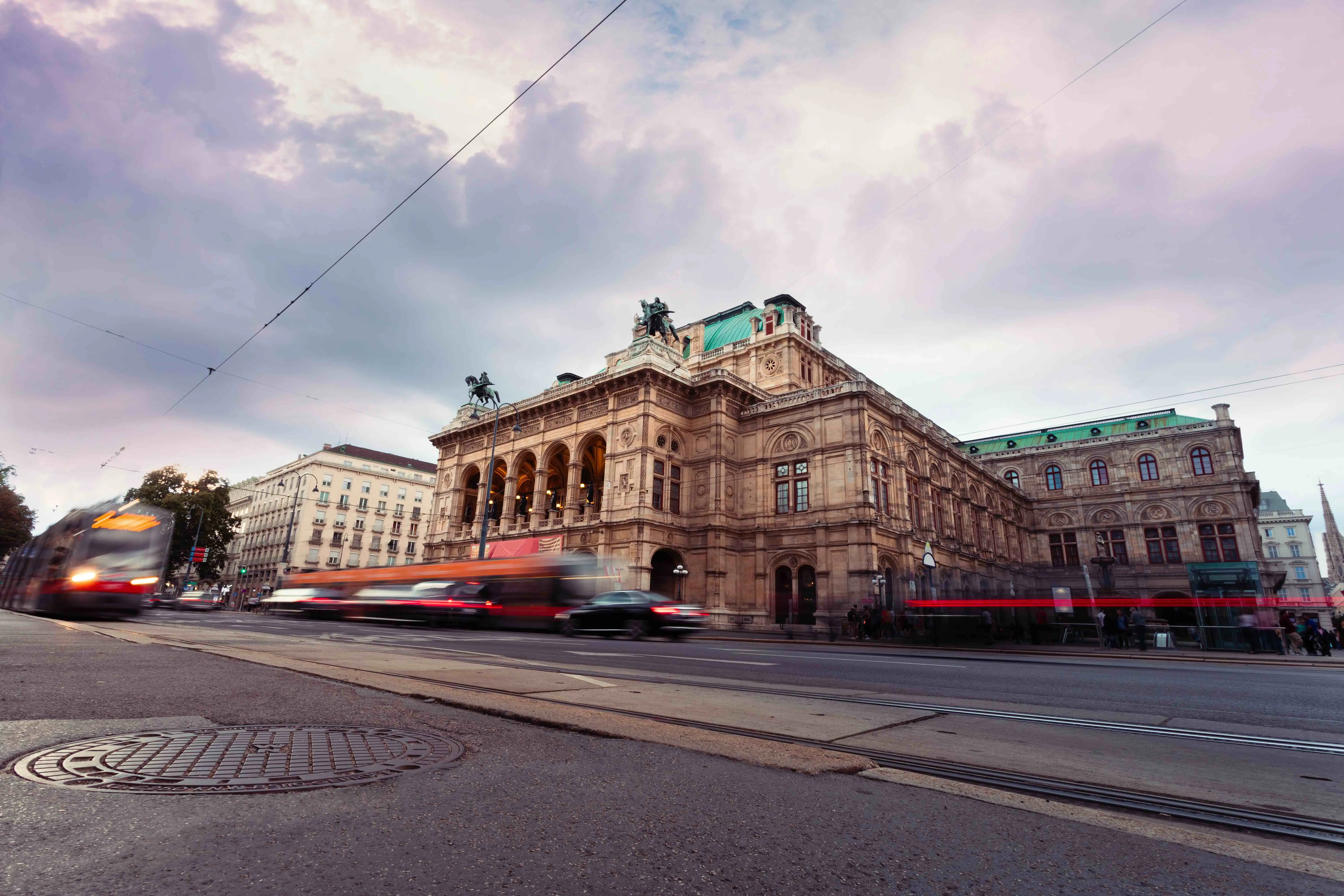 Low angle shot of Vienna Opera House, a beige coloured old-fashioned building with turquoise roofs and two statues of people on horses on either side of the front roof. Below is an action shot of vehicles moving, making them blurry, and a drain in the ground is in the left forefront. The sky is grey and cloudy.  