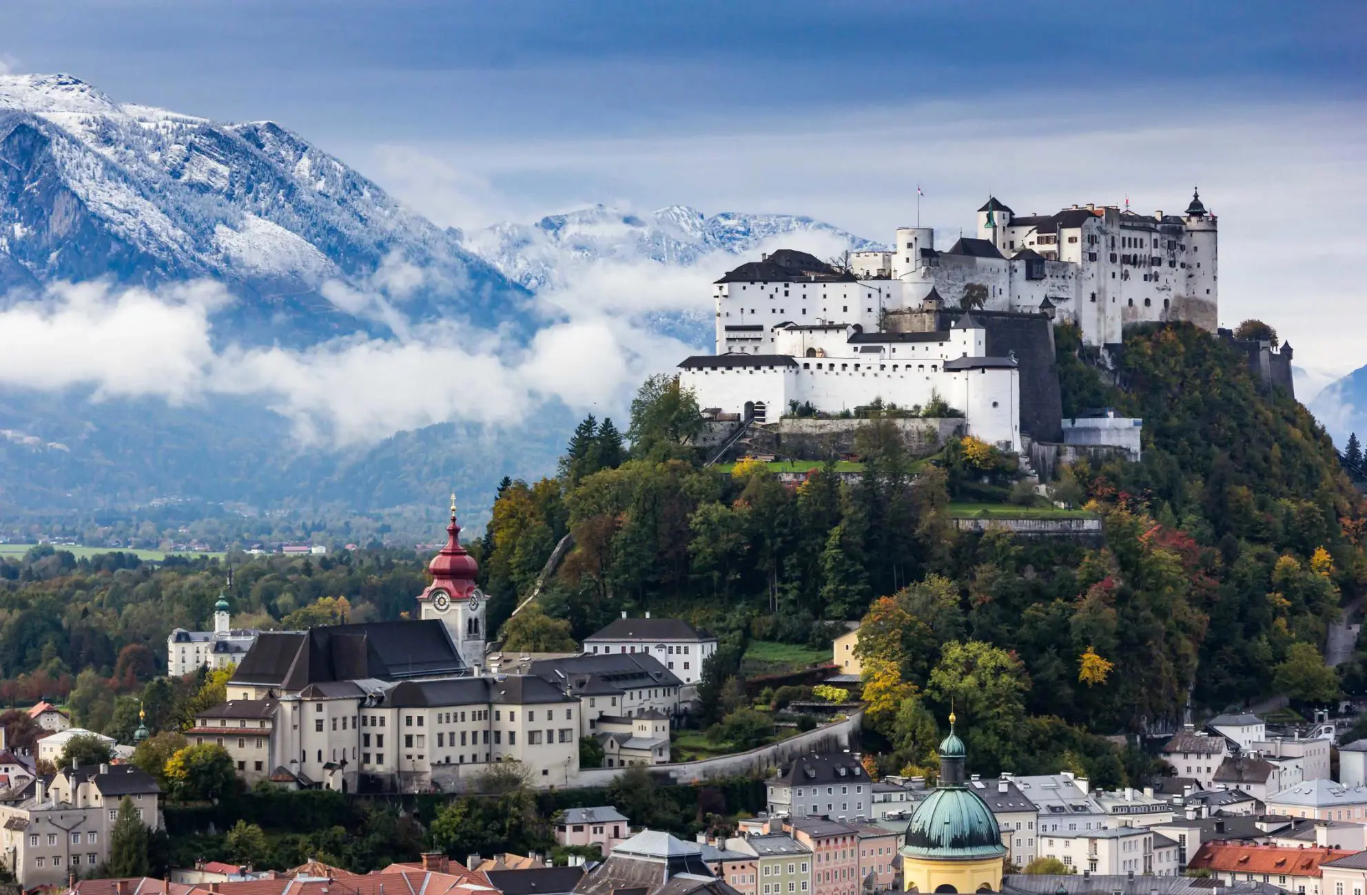 View of the Hohensalzburg Fortress, a white castle with black roofs on top of a grassy hill. At the bottom of the hill is a white large building with a red turret. Behind these is a large mountain with snow on and clouds in front of it.