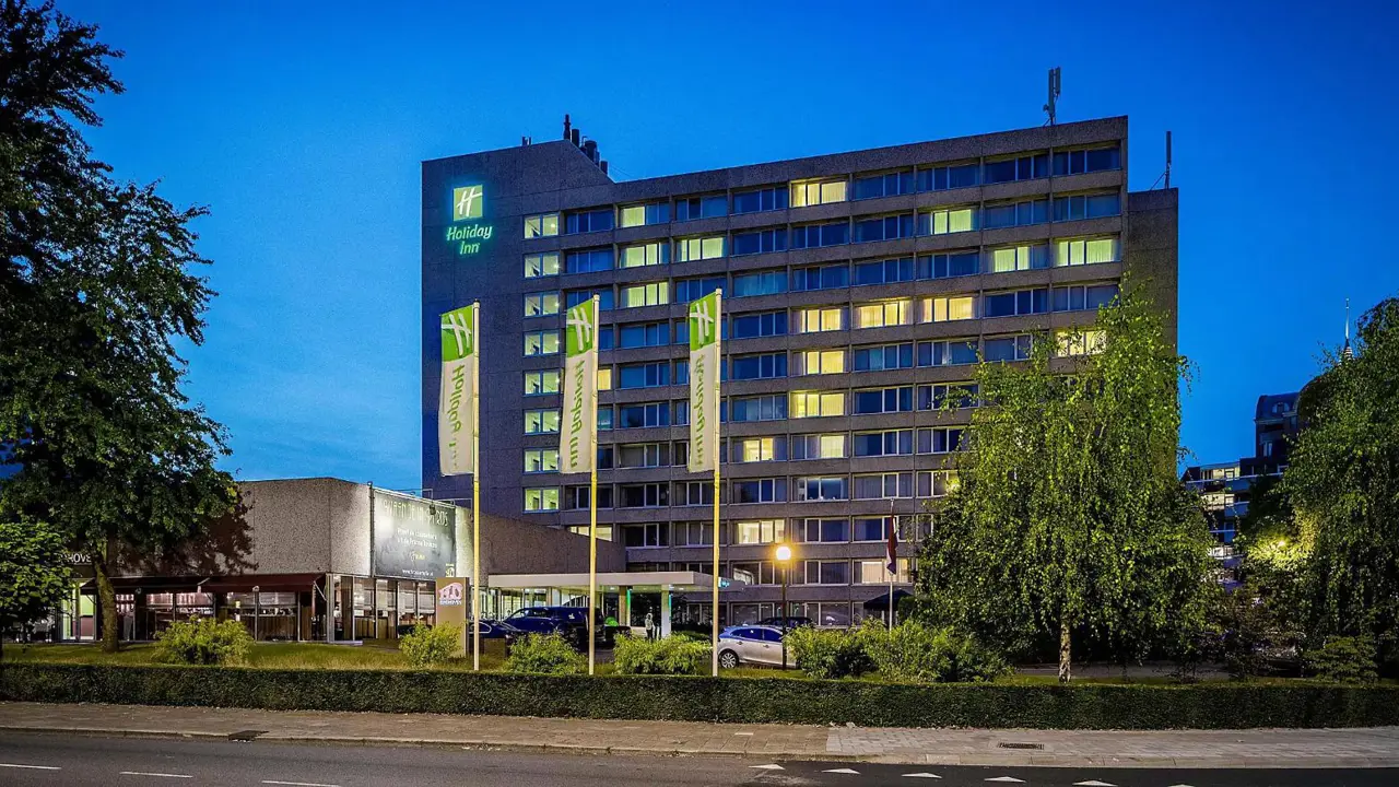 Exterior of the Holiday Inn Eindhoven, a modern building with signage on the top left, set beside a road with parked cars and green trees