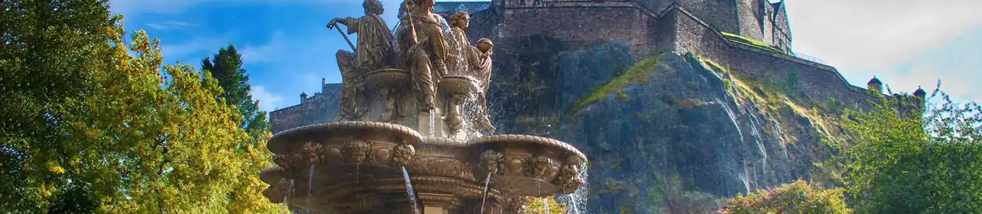 A view of Edinburgh Castle perched high on a rocky hill in the background, with a decorative fountain in the foreground below, surrounded by greenery