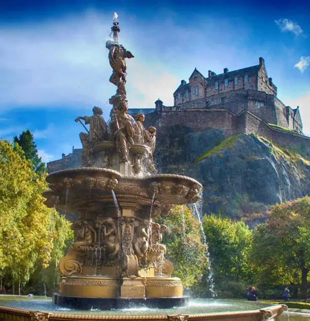 A view of Edinburgh Castle perched high on a rocky hill in the background, with a decorative fountain in the foreground below, surrounded by greenery