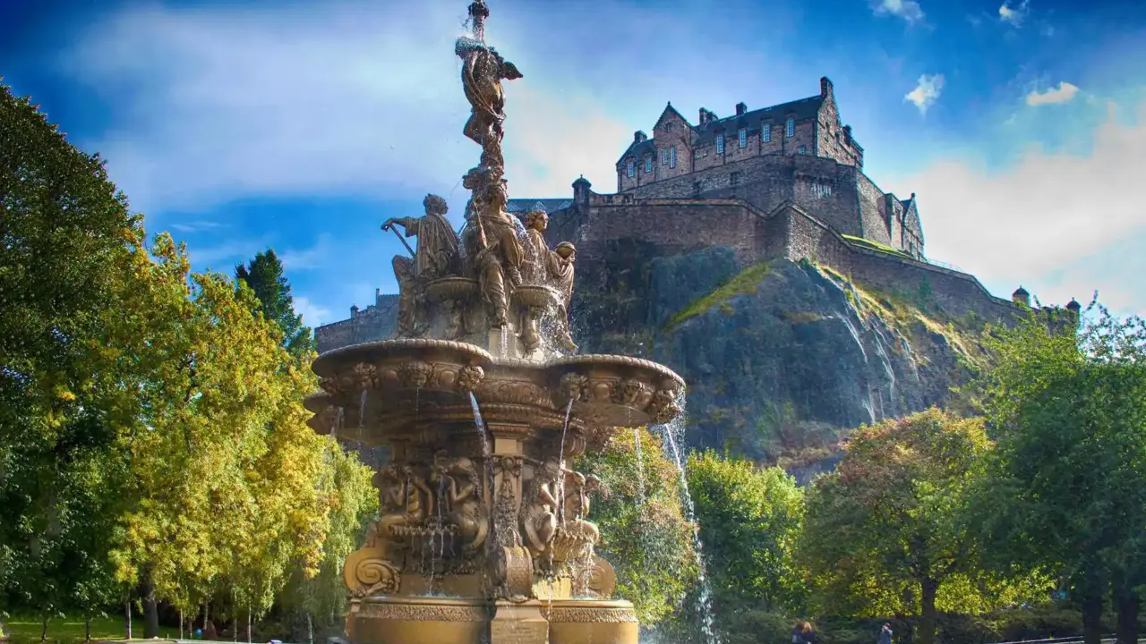 A view of Edinburgh Castle perched high on a rocky hill in the background, with a decorative fountain in the foreground below, surrounded by greenery
