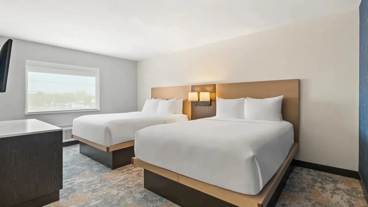 Hotel suite featuring two queen-sized beds with white bedding, a small sitting area, modern furnishings, and a large window letting in daylight