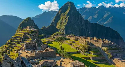 A panoramic view of Machu Picchu in Peru, showing ancient Incan ruins built into a green mountain ridge under a clear blue sky, with Huayna Picchu towering in the background