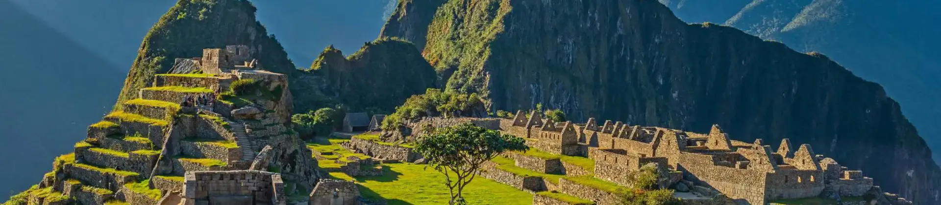 A panoramic view of Machu Picchu in Peru, showing ancient Incan ruins built into a green mountain ridge under a clear blue sky, with Huayna Picchu towering in the background
