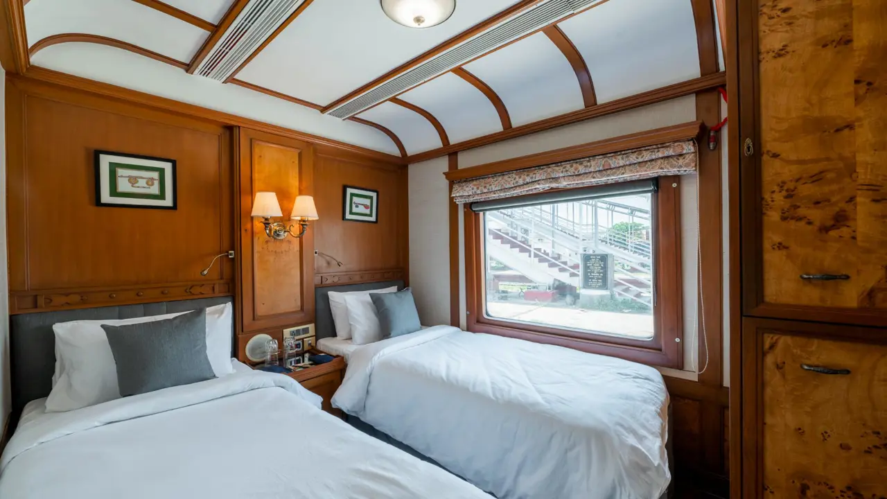 Interior of a Deccan Odyssey twin room, showing two neatly made beds, elegant wooden furnishings, and a large window with a patterned blind