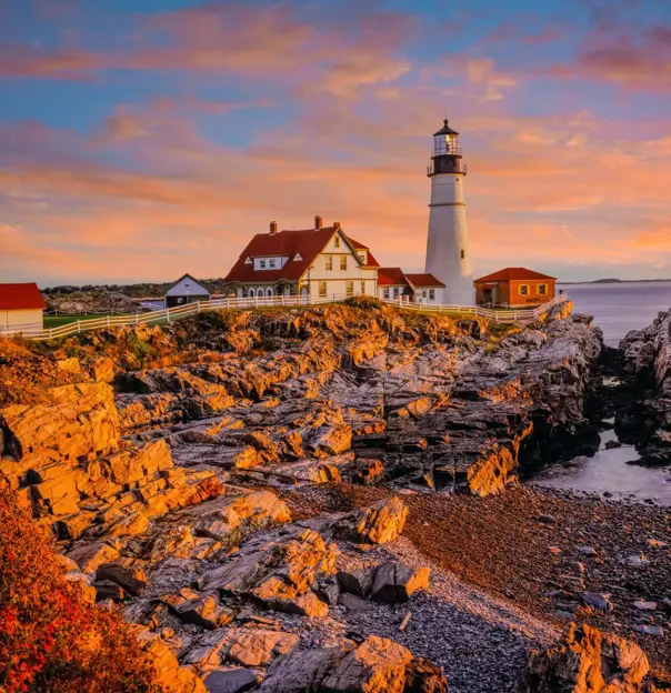 Portland Head Lighthouse on the rocky Maine coast at sunset, with warm colours in the sky reflecting over the Atlantic Ocean