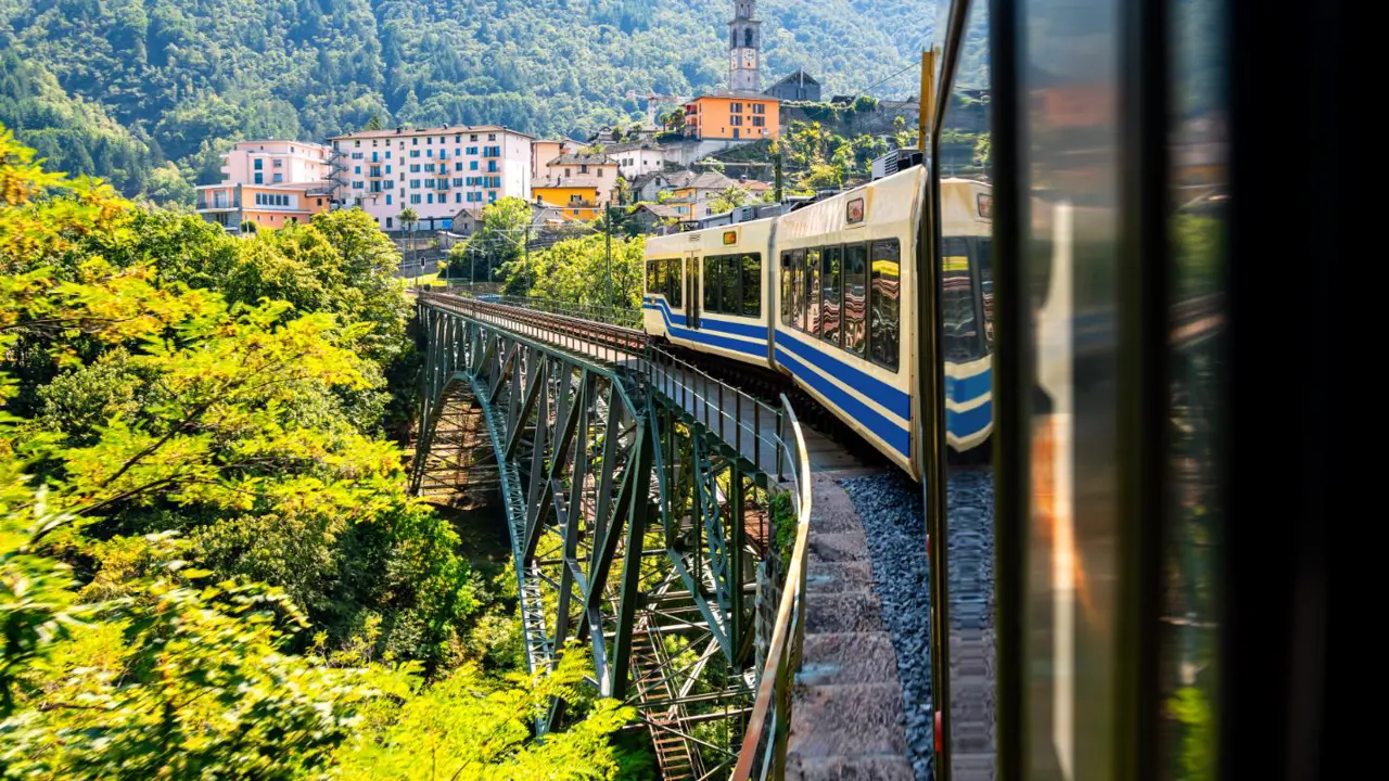 Centovalli Railway bridge in Switzerland with a train crossing, surrounded by mountainous countryside, and a town visible in the distance