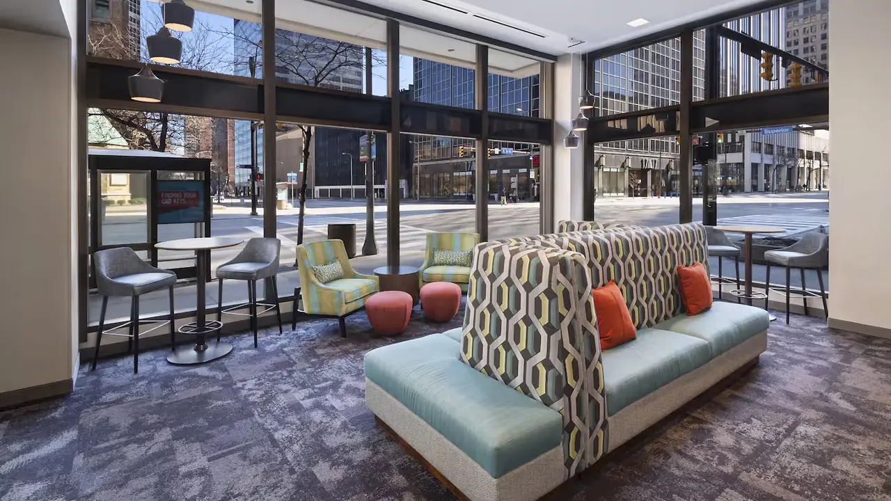 The lounge at Hampton Inn Downtown, Cleveland, featuring contemporary seating in the centre with green upholstery and deep orange cushions. There are large street-facing windows that fill the space with natural light