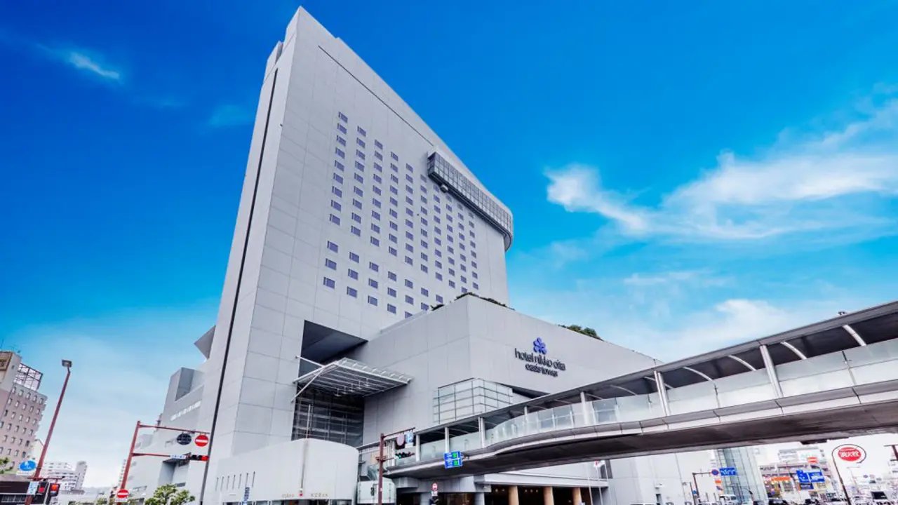 Exterior of Hotel Nikko Ōita Oasis Tower, a tall, modern building with a skybridge connecting to the main entrance and a bright blue sky overhead