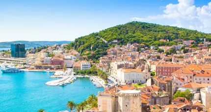 A panoramic view of Split, Croatia, showing the turquoise Adriatic Sea, a bustling harbour with yachts, and the terracotta rooftops of Old Town at the foot of lush green Marjan Hill