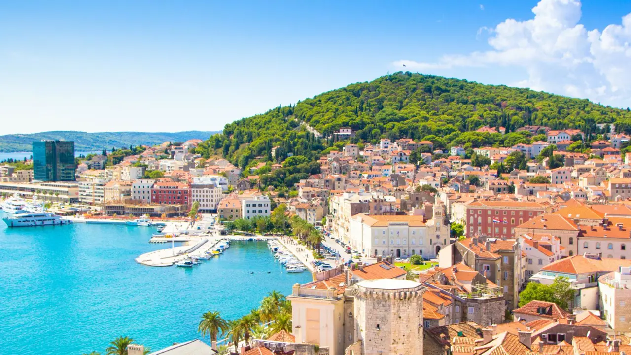 A panoramic view of Split, Croatia, showing the turquoise Adriatic Sea, a bustling harbour with yachts, and the terracotta rooftops of Old Town at the foot of lush green Marjan Hill