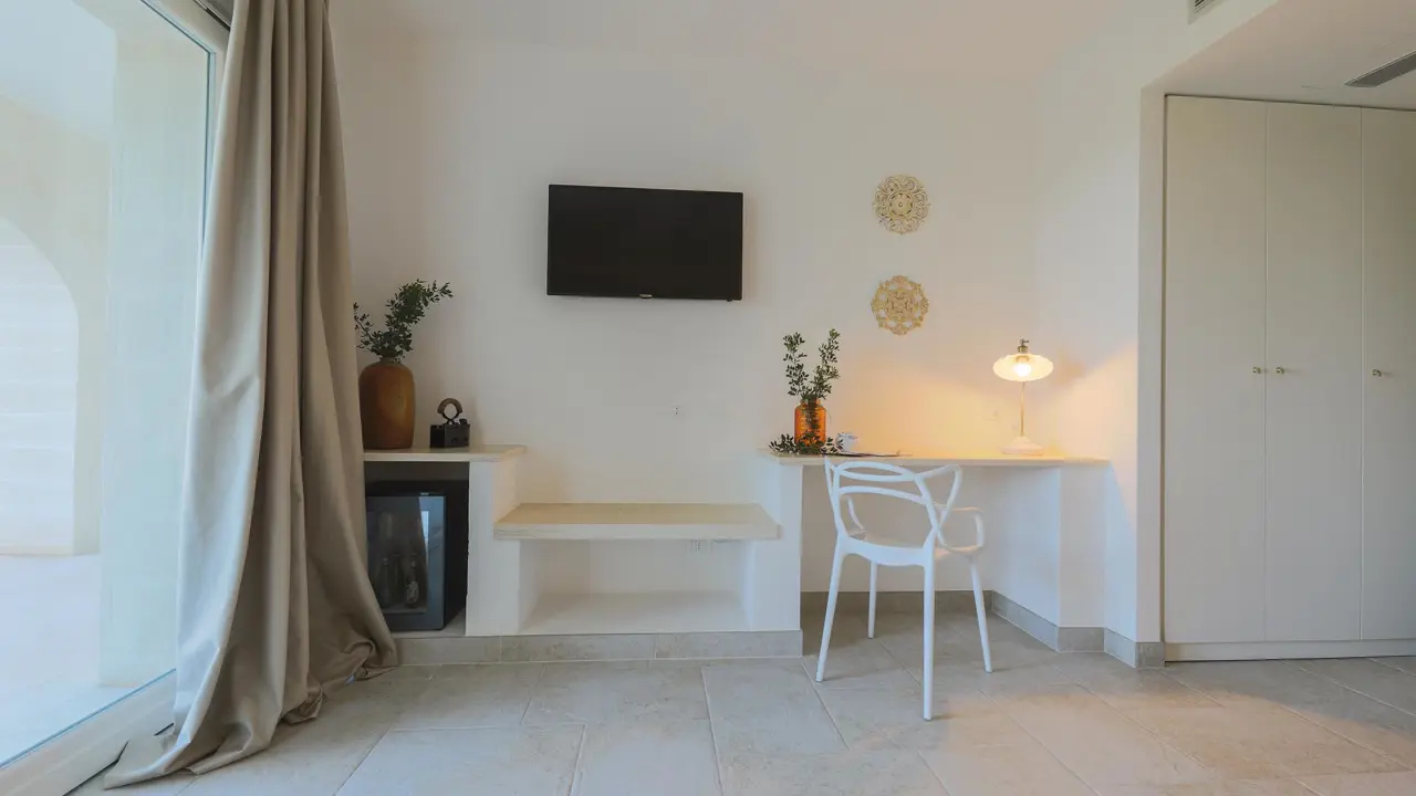 Bright minimalist room at Masseria Casselli Hotel featuring a wall-mounted TV, white desk and chair, a table lamp, decorative plants, and beige curtains covering a large glass door