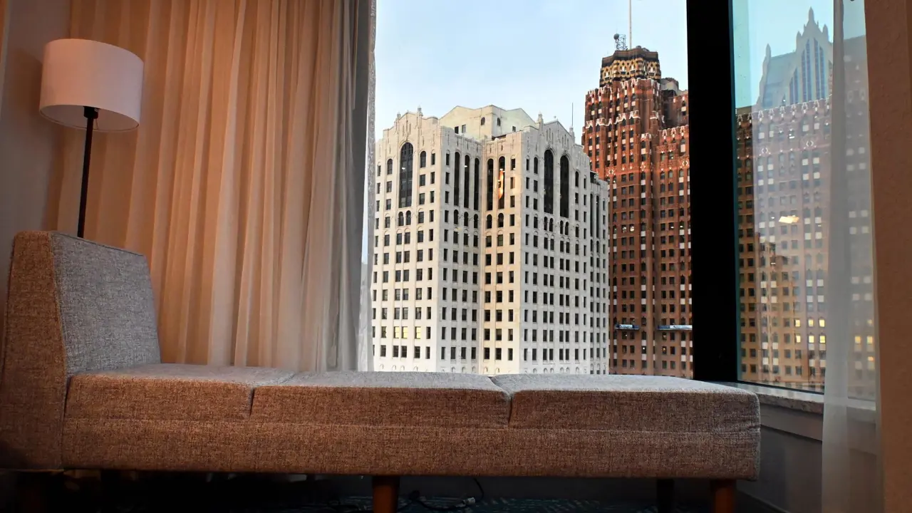 City view from a standard room at Fort Pontchartrain hotel, featuring a modern grey chaise lounge, floor lamp, and large windows overlooking historic high-rise buildings in downtown Detroit