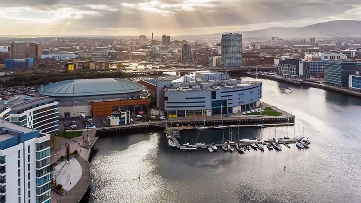 High angle view of Ulster Museum, a spherical, grey building on the edge of the River Lagan and next to a harbour with parked boats. The rest of the city of Belfast can be seen below a cloudy sky