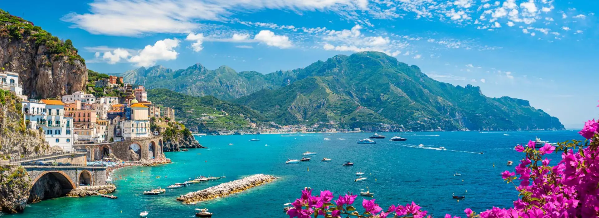 Scenic View Of Amalfi Coast Italy, showing the sea and boats, mountain in the distance, town on the edge of a cliff and pink flowers in the forefront