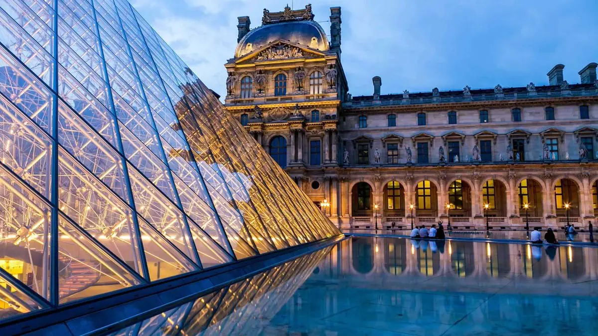 Top of a old-fashioned building with many windows and archways, to the left, a modern, glass roof with a cross design