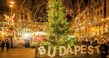 A square in Budapest at Christmas, in the front, a 'Budapest' sign, with a Christmas tree with lights and red baubles behind. Behind this, lit up decorations with hanging baubles, strung across the square.