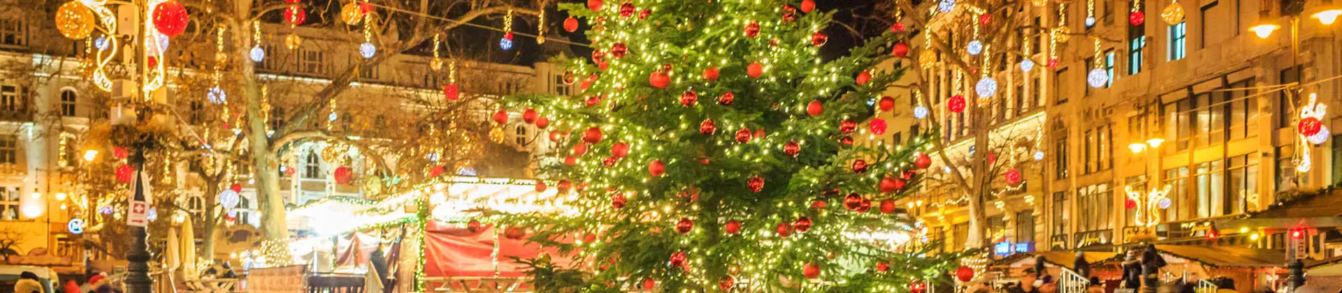 A square in Budapest at Christmas, in the front, a 'Budapest' sign, with a Christmas tree with lights and red baubles behind. Behind this, lit up decorations with hanging baubles, strung across the square.