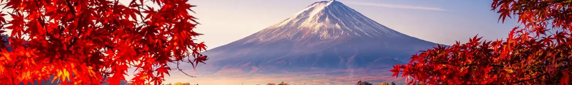 Mount Fuji reflected in Lake Kawaguchi, framed by vivid red autumn leaves, with a sunlit lakeside town between the water and the mountain