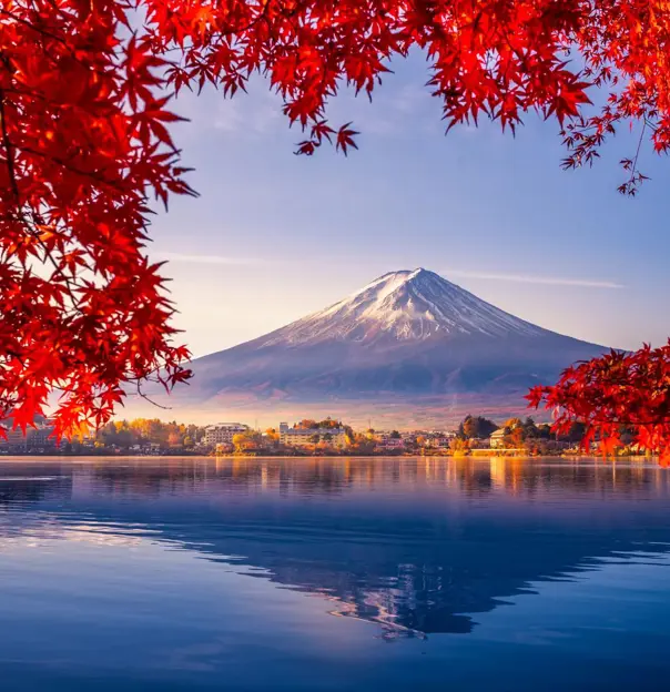 Mount Fuji reflected in Lake Kawaguchi, framed by vivid red autumn leaves, with a sunlit lakeside town between the water and the mountain