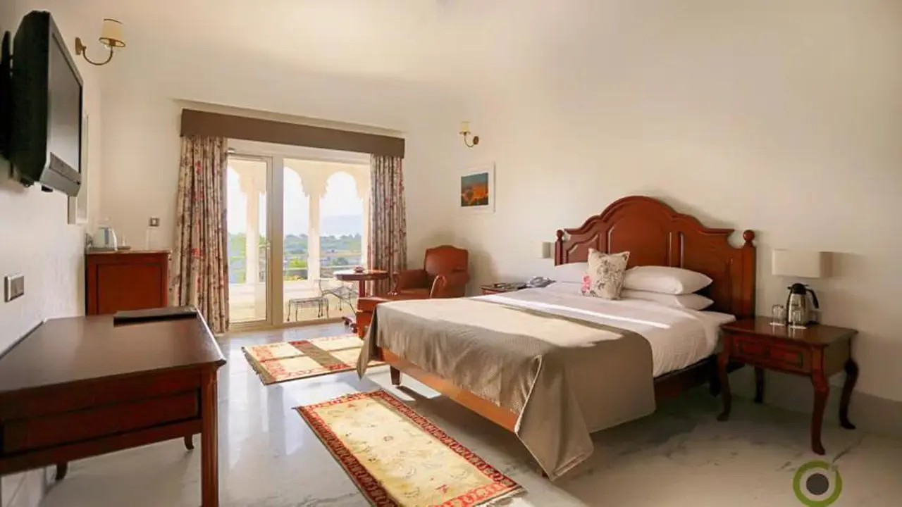 Spacious guest room at Fateh Niwas Hotel, Udaipur, with a large bed, classic wooden furniture, and a balcony view of the lake