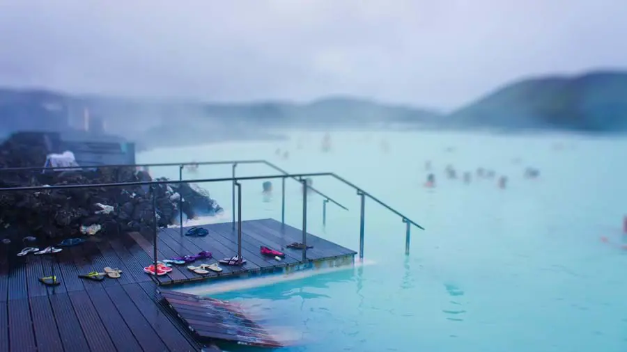 Geothermal lagoon in Iceland - bright blue water with steam coming off, mist clouding the view. In the forefront, a wooden platform descending into the water, with pairs of flip flops. 