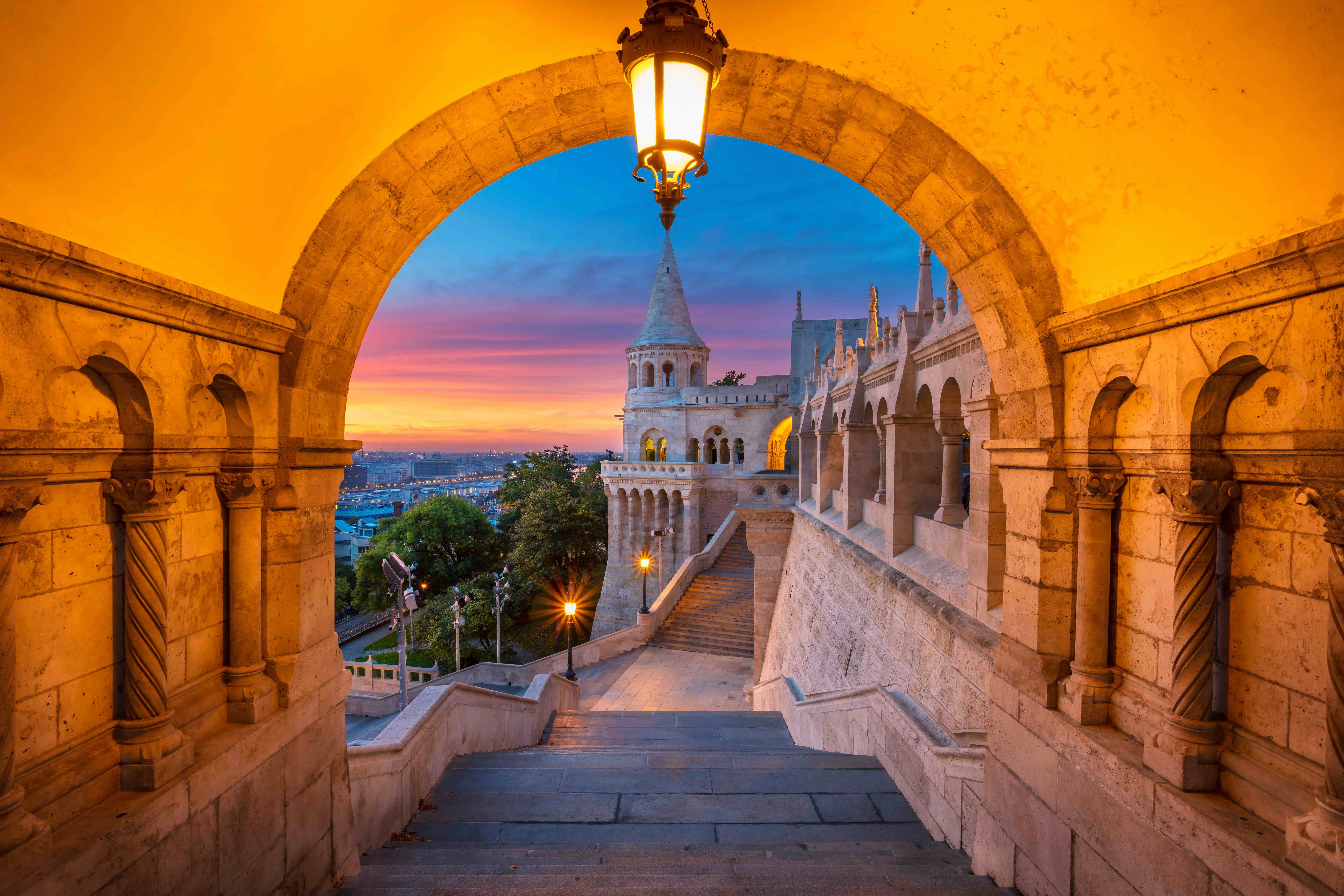 View of the side of Buda Castle in Budapest from a warmly lit archway, showing a grey turret in front of blue, pink and orange sky