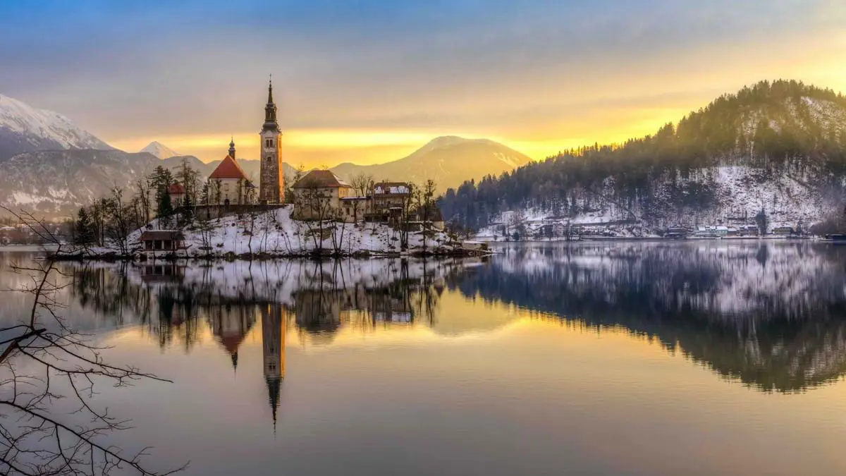 A lake at winter time, with a church in the centre with a slim tower and spiked turret. Mountains in the distance