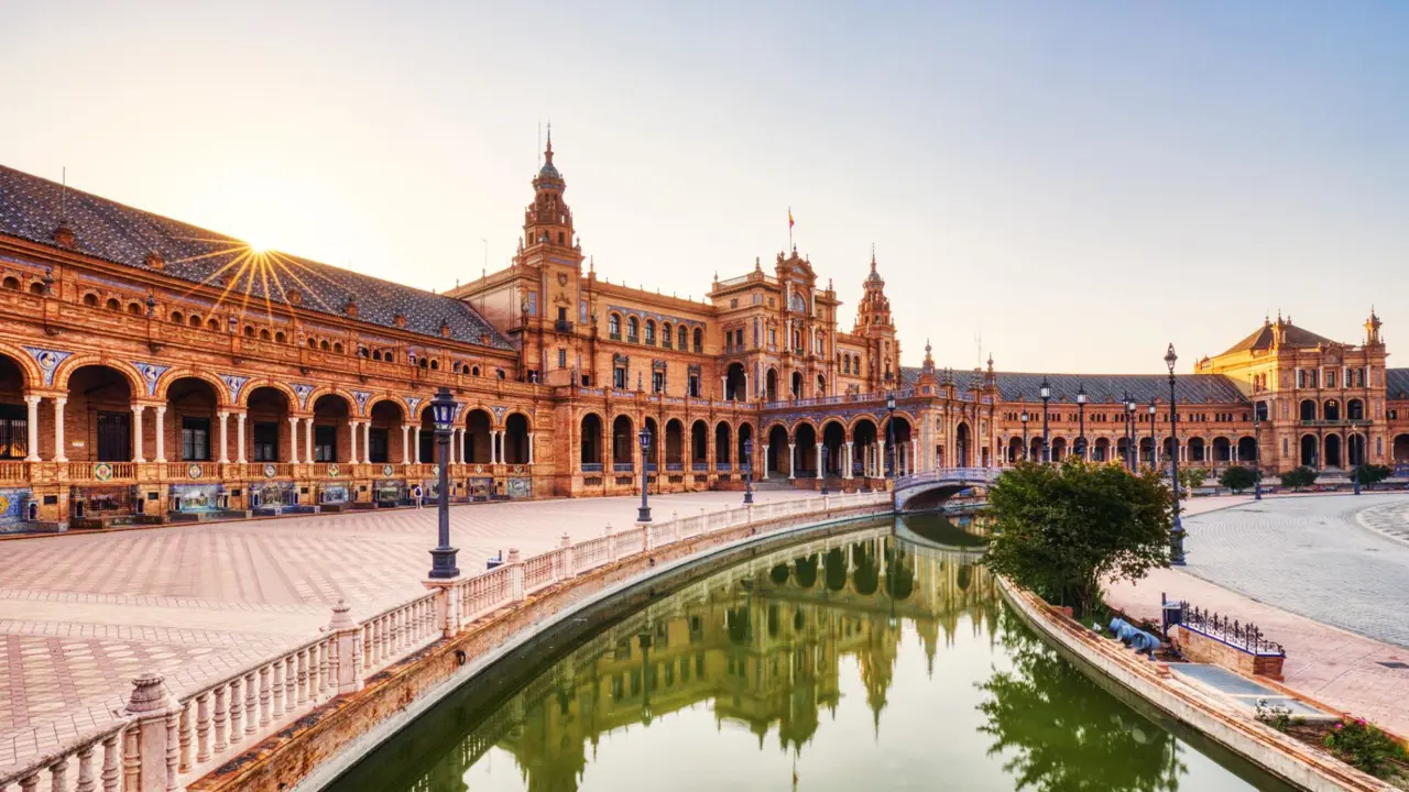 Wide view of Plaza de España in Seville, featuring its grand semicircular building, ornate tilework, and the small river with a bridge crossing it in the distance