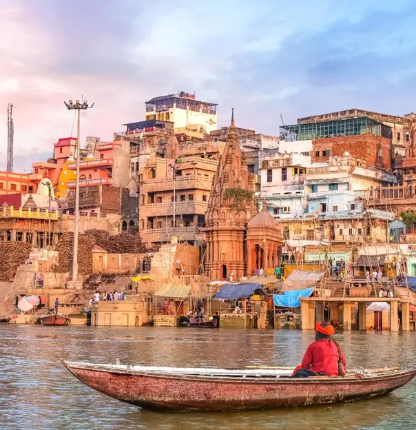 A boatman rowing on the River Ganges in Varanasi, facing the colourful temples and crowded ghats along the ancient city’s waterfront under a soft pastel sky