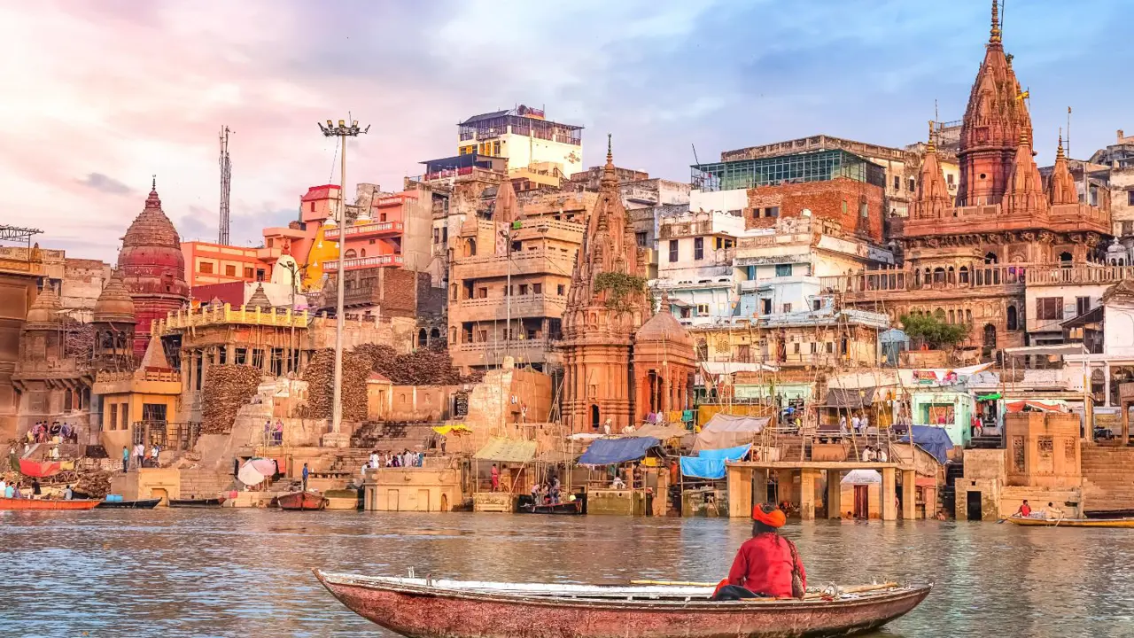 A boatman rowing on the River Ganges in Varanasi, facing the colourful temples and crowded ghats along the ancient city’s waterfront under a soft pastel sky