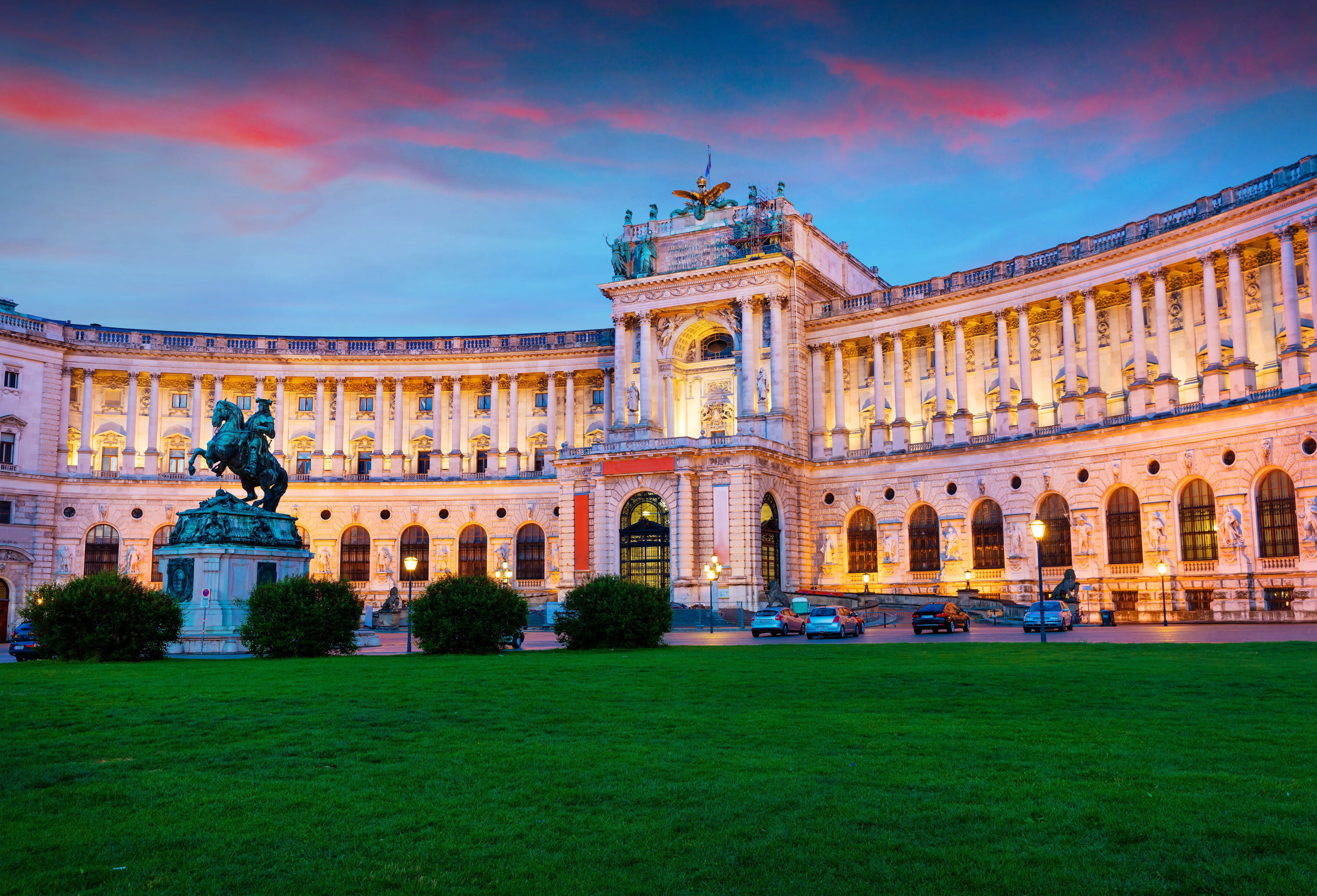Low angle shot of a cream coloured, wide, old-fashioned building. In front is a green statue of a man riding a horse, next to a dark green lawn and four circular bushes.