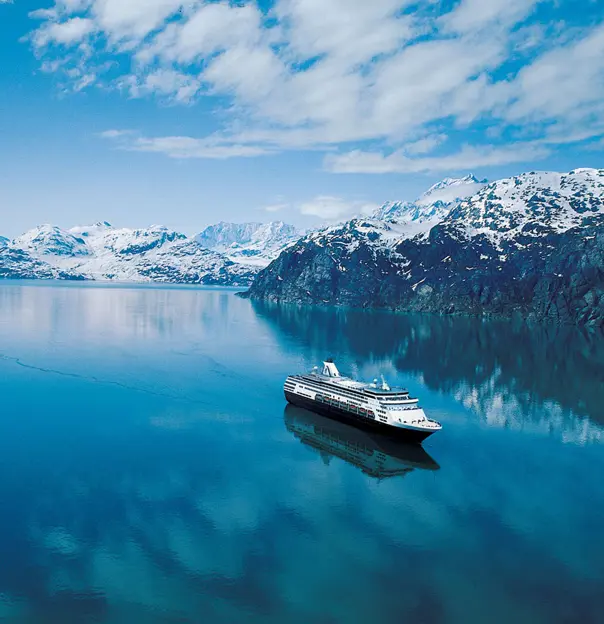A Holland America Line cruise ship sailing through the clear blue waters of the Inside Passage near Alaska, with snow-capped mountains in the distance