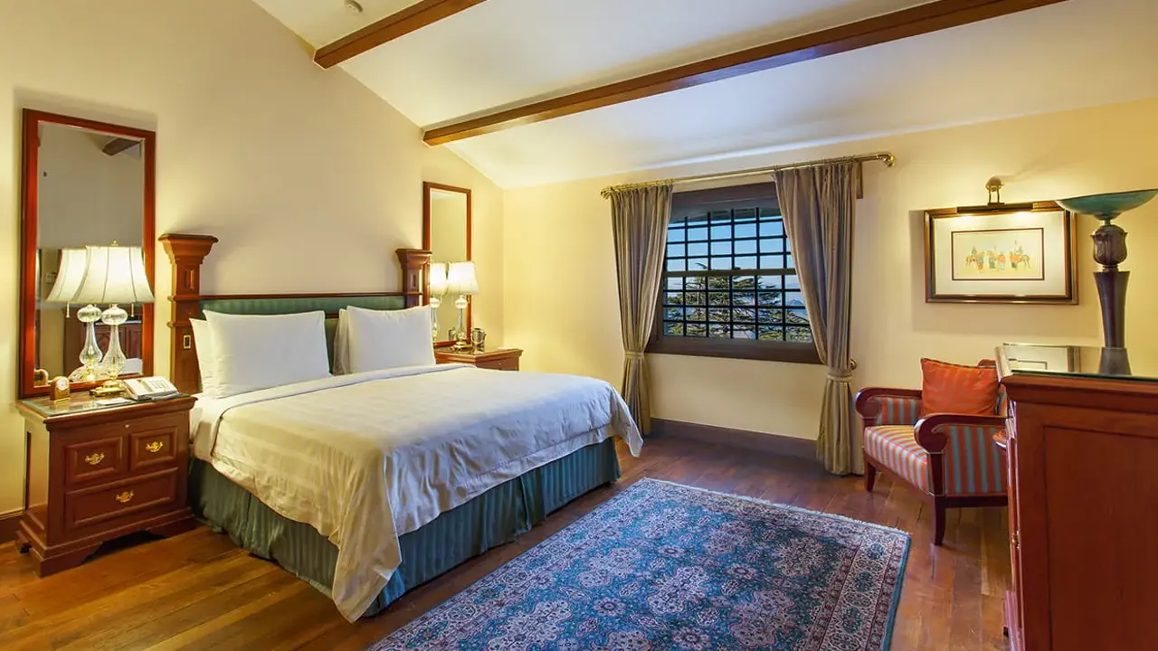 A double room at the Oberoi Cecil hotel in Shimla, featuring wooden floors, a blue patterned rug, cosy warm furnishings, and a window with views of the surrounding trees
