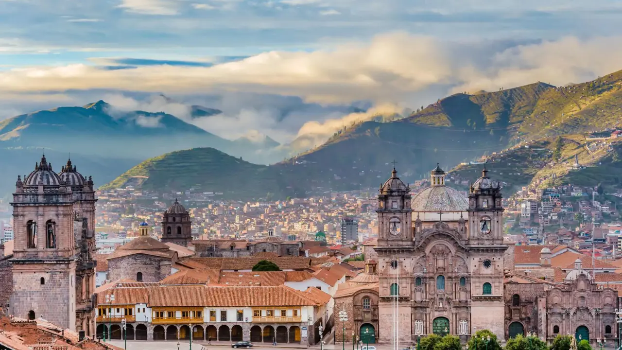A panoramic view of Cusco, Peru, showcasing historic colonial buildings with terracotta roofs against a backdrop of green mountains and a cloudy sky.A panoramic view of Cusco, Peru, showcasing historic colonial buildings with terracotta roofs against a backdrop of green mountains and a cloudy sky