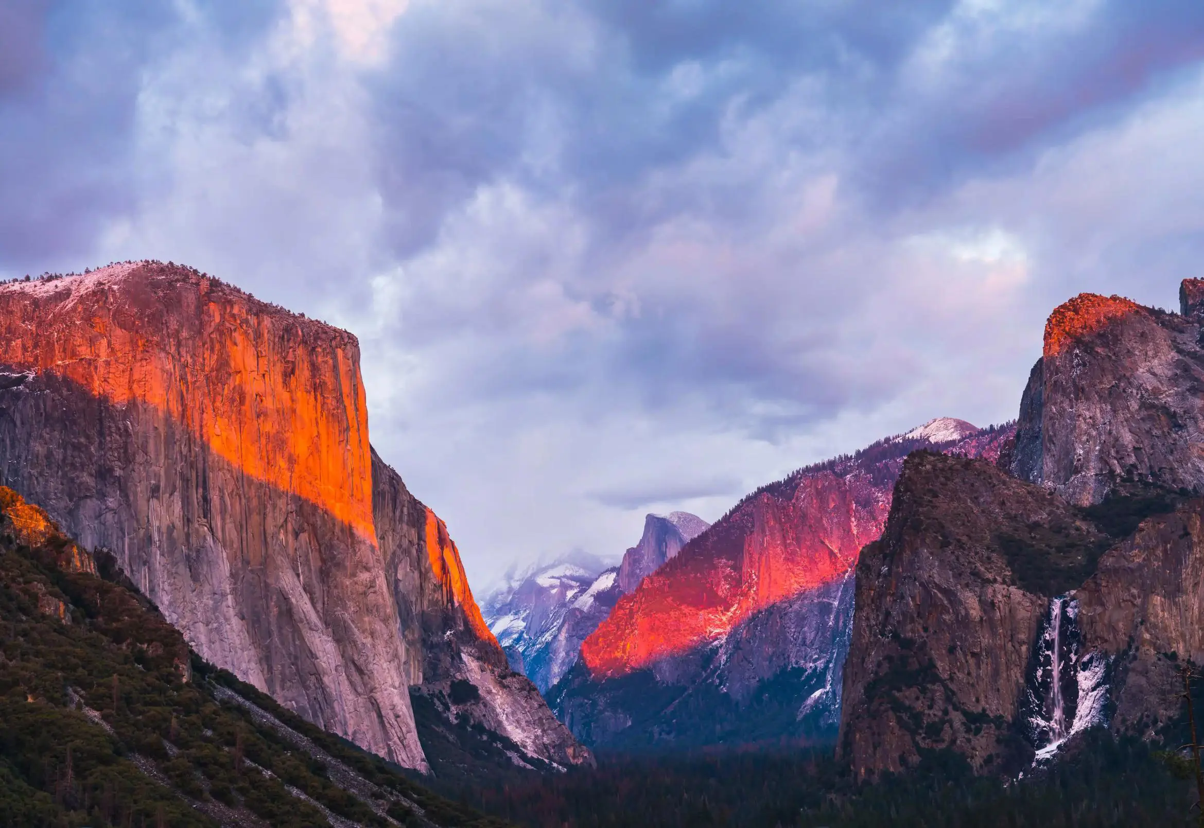 A dramatic view of Yosemite’s El Capitan and surrounding mountains bathed in warm, glowing sunset light under a cloudy sky