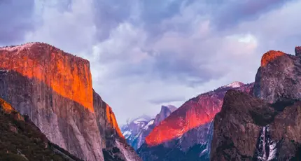 A dramatic view of Yosemite’s El Capitan and surrounding mountains bathed in warm, glowing sunset light under a cloudy sky