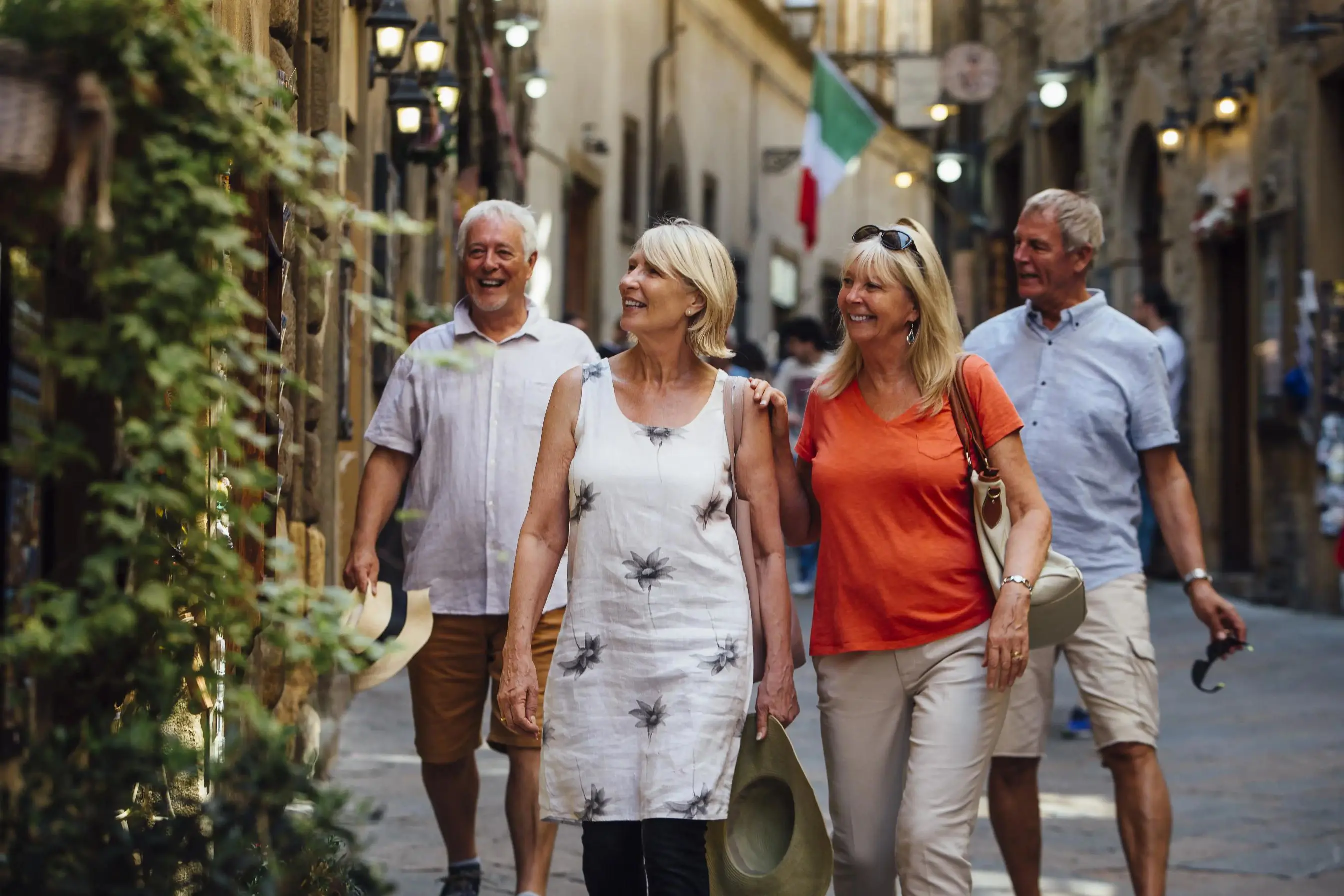 Smiling elderly friends stroll through a charming Italian street with historic buildings and an Italian flag in the background