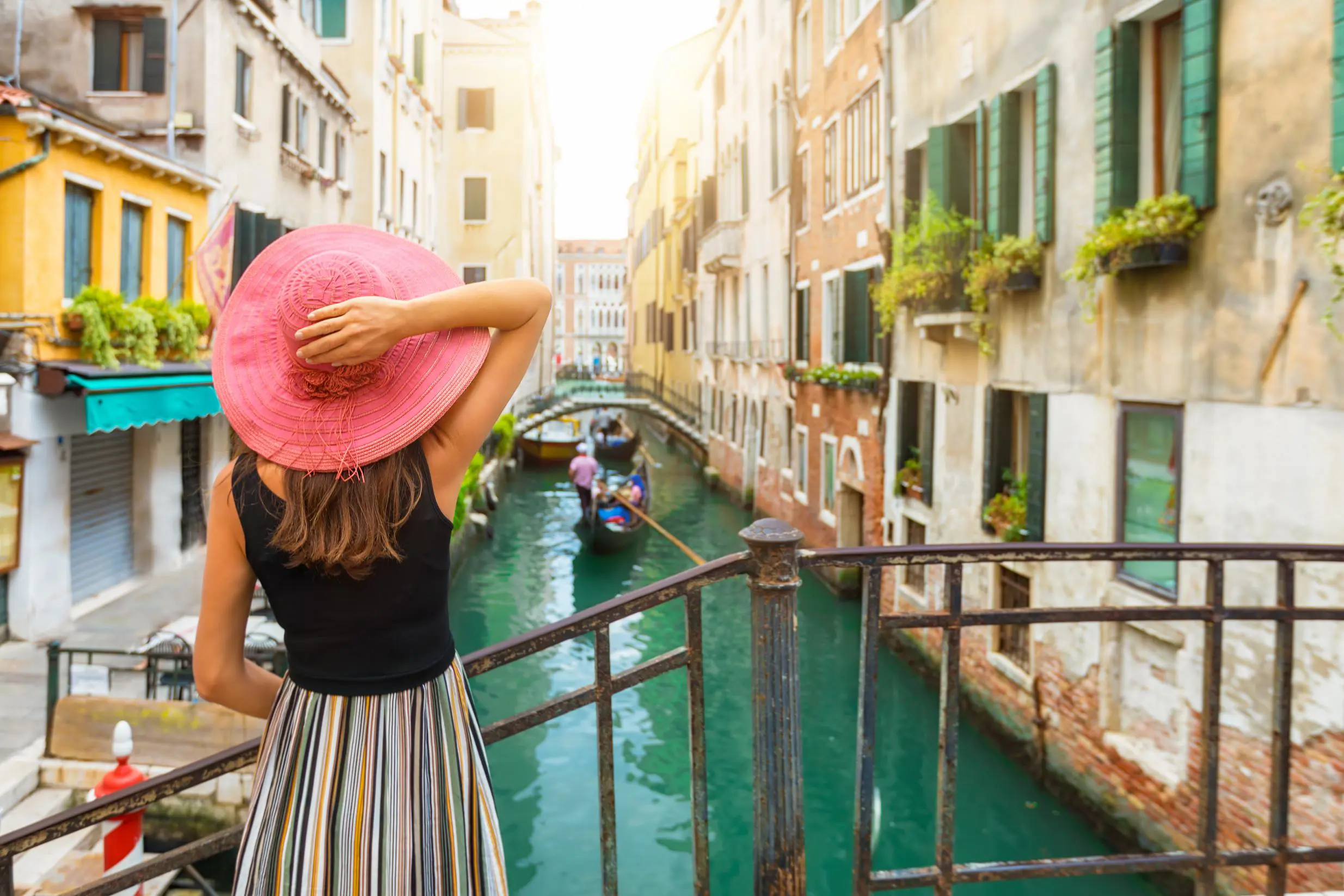A woman in a striped skirt and pink hat standing on a bridge, looking out over a canal with gondolas in Venice, Italy, on a sunny day
