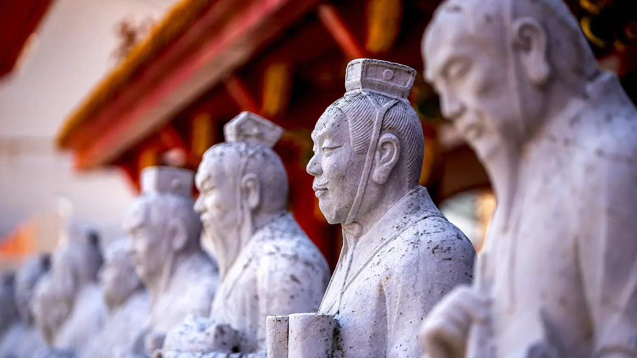Stone statues of ancient scholars holding scrolls at the Confucius Temple in Nagasaki, Japan, with a red-roofed structure in the background